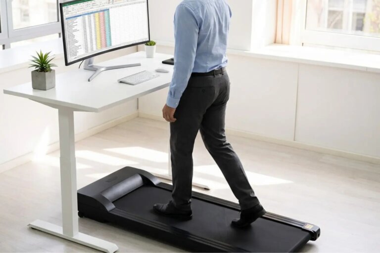 Anonymous man in smart casual attire walks on a desk treadmill while working at a standing desk with a monitor and plant.